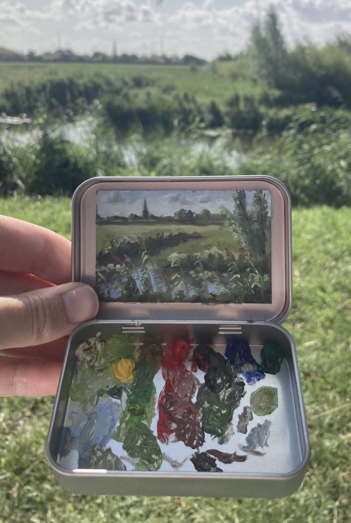 Blikvanger miniatuur schilderij in blikje met bijbehorende originele palet van zicht op Voorhout met water bomen en een kerk zichtbaar in de verte