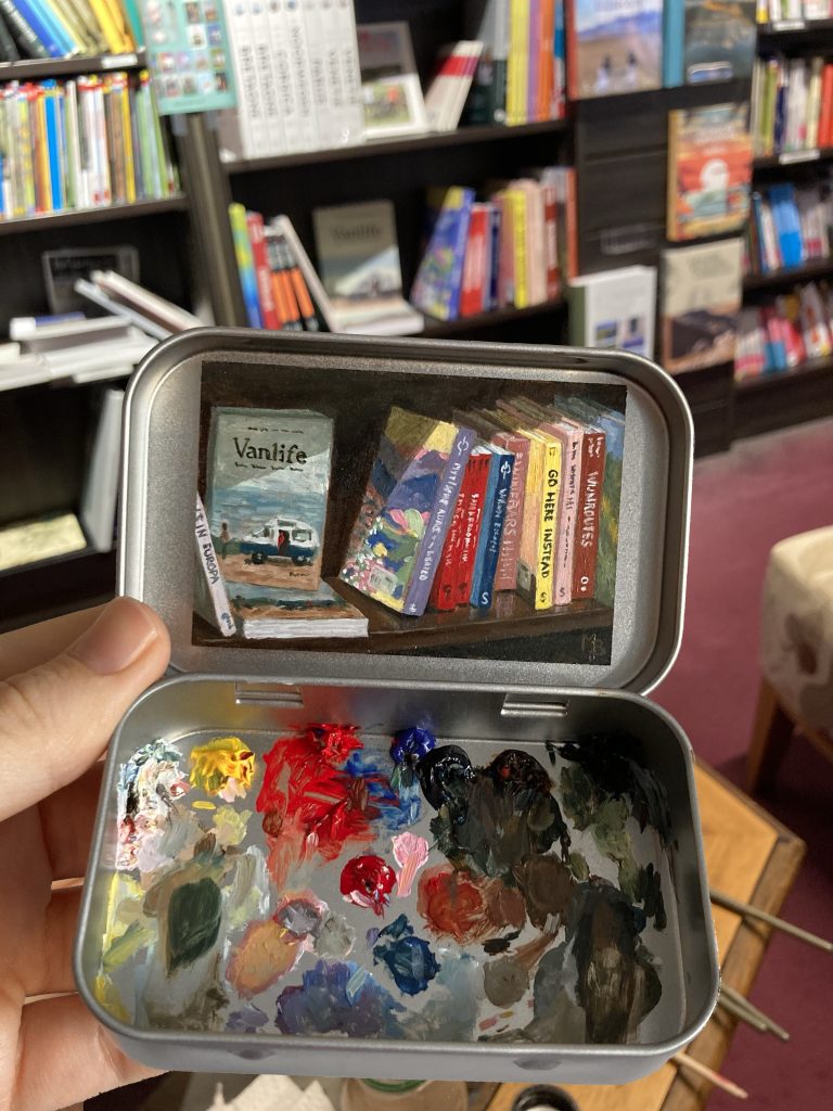 Blikvanger miniatuur schilderij in blikje met bijbehorende originele palet van reisboeken op een boekenplank in boekhandel van der Meer Noordwijk