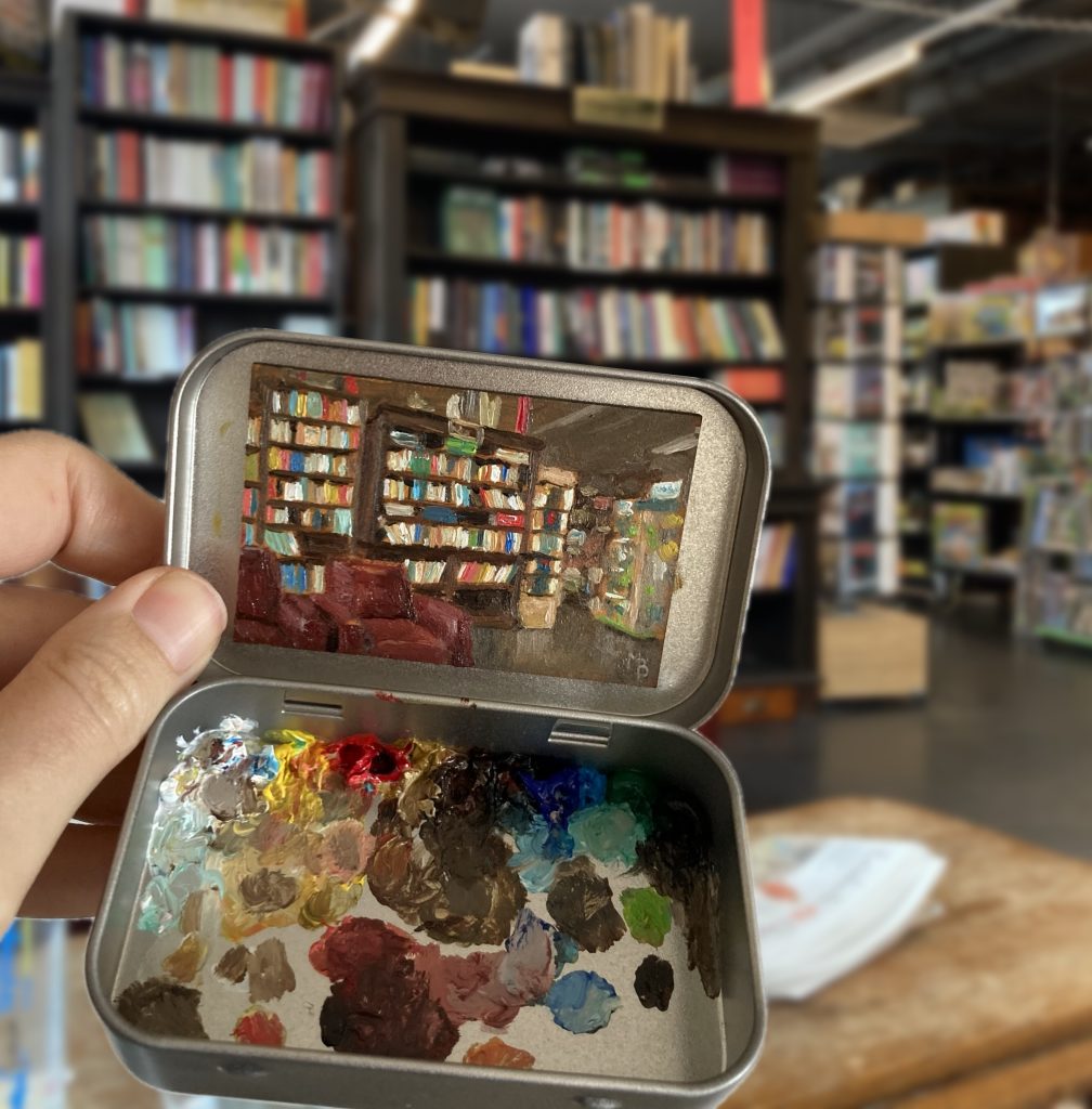 Blikvanger miniatuur schilderij in blikje met bijbehorende originele palet van boekhandel van der meer met interieur winkel met boekenkasten op de achtergrond