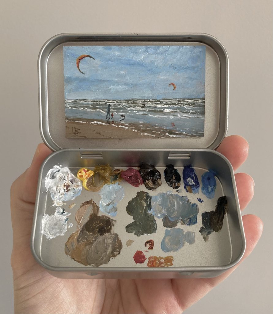 Blikvanger miniatuur schilderij in blikje met bijbehorende originele palet van zee en strand op winderige dag met kitesurfers en ouder en kind met hond die op strand lopen